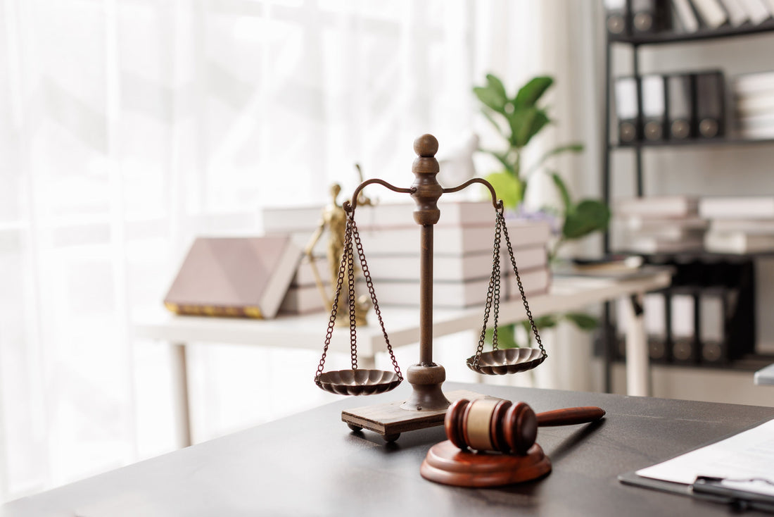 Waage und Richterhammer auf einem Schreibtisch, im Hintergrund Justitia-Figur, Gesetzbücher und Aktenordner