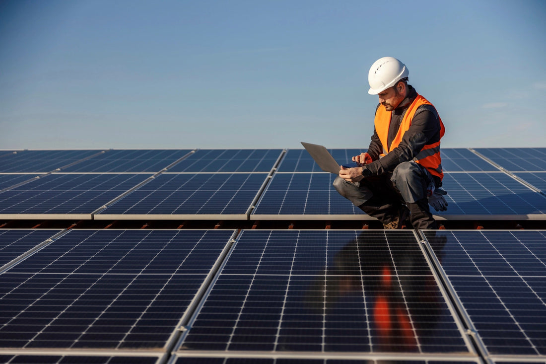 Techniker mit Schutzhelm und Warnweste kniet zwischen Solarmodulen und arbeitet an einem Laptop