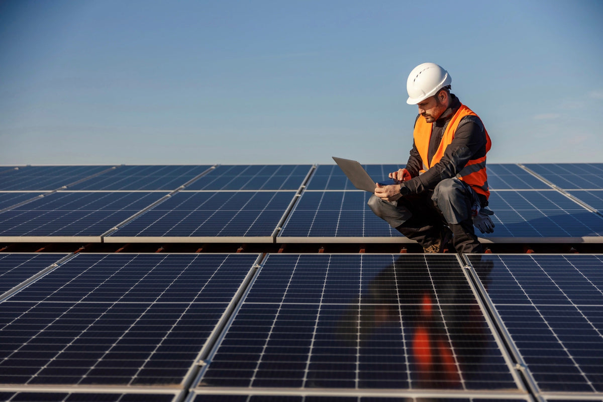 Techniker mit Schutzhelm und Warnweste kniet zwischen Solarmodulen und arbeitet an einem Laptop
