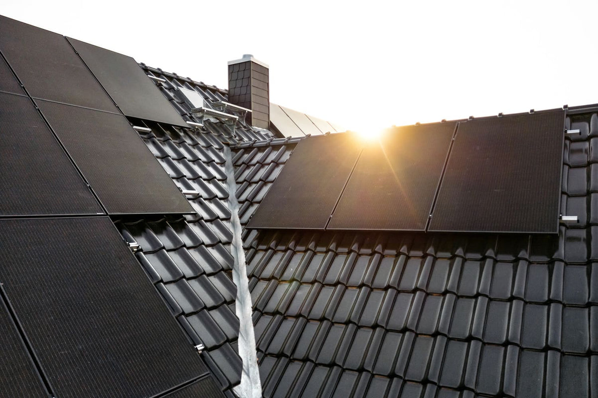 Schwarze Solarmodule auf einem dunklen Ziegeldach mit Schornstein, im Hintergrund geht die Sonne hinter dem Dachfirst auf
